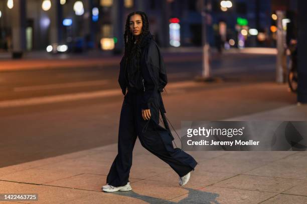 Guest is seen wearing black oversize nylon bomber jacket, matching black nylon parachute pants, gold and pearl necklaces and white Adidas Astir...