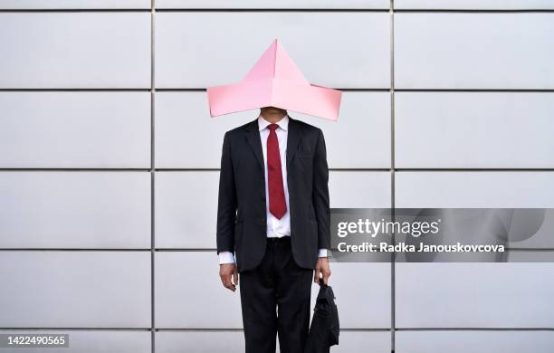 businessman in black suit standing with a laptop bag with a funny pink paper hat over his head - estancado fotografías e imágenes de stock