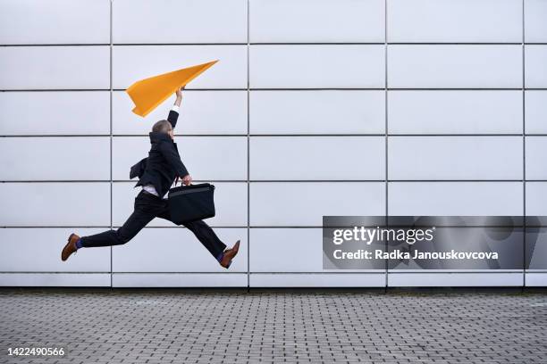 businessman in black suit running fast and jumping with a paper plane and a laptop bag - lanzamiento publicitario fotografías e imágenes de stock