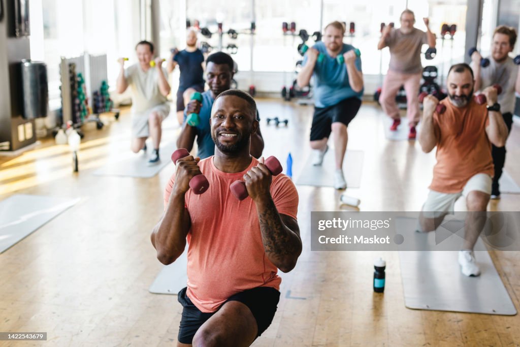 Smiling male fitness instructor practicing dumbbell exercise with men in gym