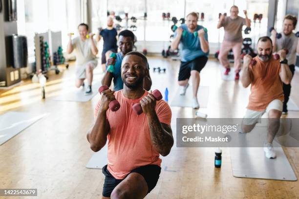 smiling male fitness instructor practicing dumbbell exercise with men in gym - grupo de hombres fotografías e imágenes de stock