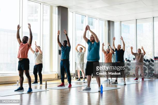 male personal trainer practicing hands raised exercise with men during group training at health club - senior people training imagens e fotografias de stock