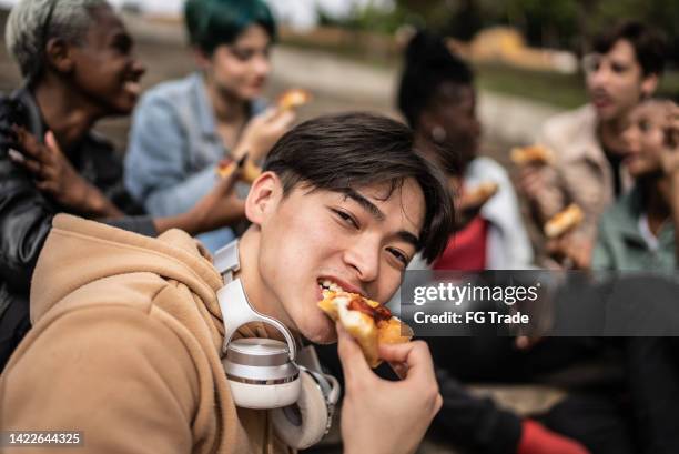 portrait of a young man eating pizza with friends in the park - social bite stock pictures, royalty-free photos & images