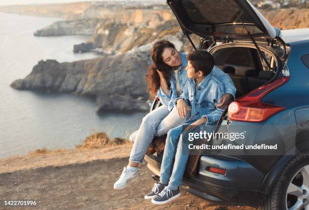 Family trip to the mountains by car. Mom is having fun with her teenage son, sitting in the trunk of a car and enjoying the view together