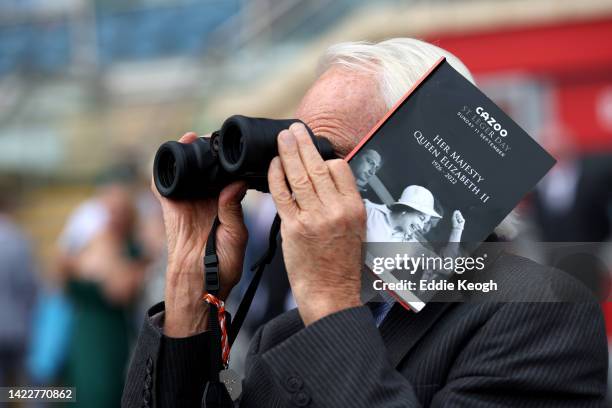 Spectator watches the racing through binoculars while holding a race program which pays tribute to Her Majesty Queen Elizabeth II, who died away at...