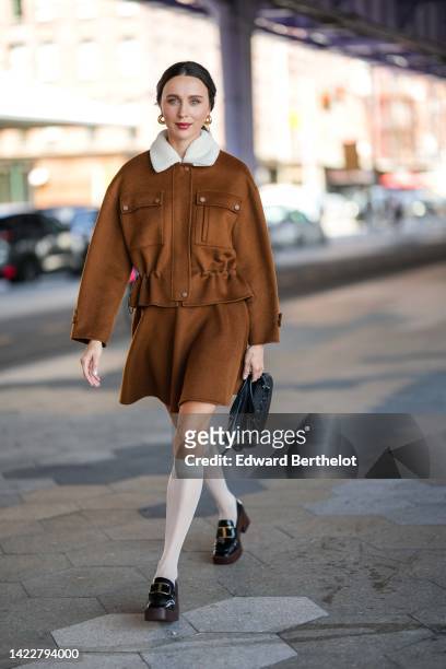Mary Leest wears gold earrings, a brown felt with large white sheep collar / cargo jacket, a matching brown felt short skirt, a black shiny leather...