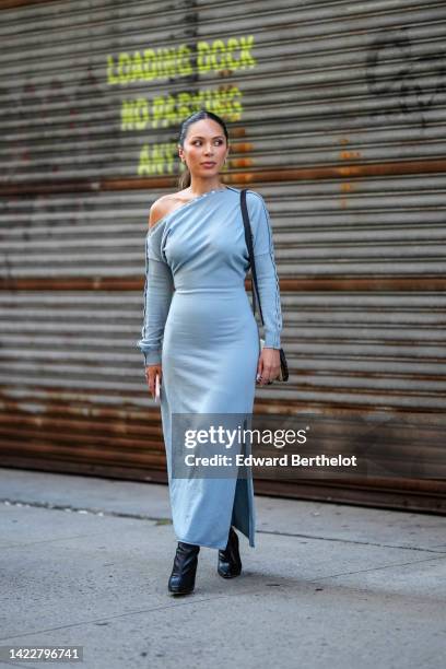 Marianna Hewitt wears silver earrings, a pale gray asymmetric shoulder / long sleeves long dress, a black shiny leather strap and beige / brown suede...