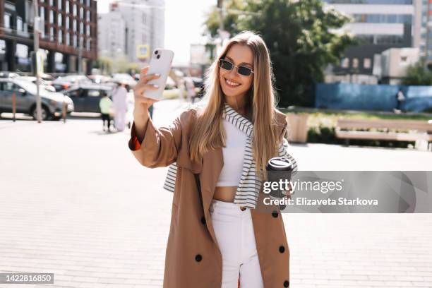 portrait of a smiling young blonde woman in sunglasses and fashionable clothes walking through the streets of the city holding a glass drinking hot coffee outdoors. a beautiful millennial student girl communicates using a mobile phone and wireless technol - holding sunglasses photos et images de collection
