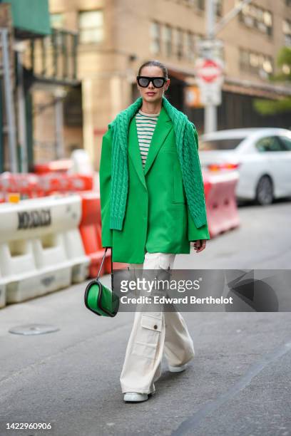 Guest wears black sunglasses, a beige and green striped print pattern t-shirt, a green oversized blazer jacket, a green braided wool pullover, white...