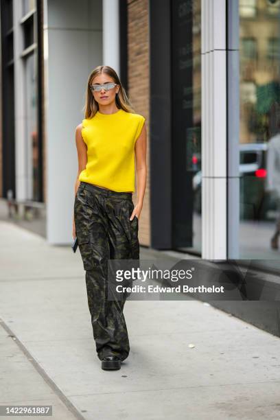 Guest wears gray futurist sunglasses, a yellow short sleeves wool pullover, black and khaki military print pattern wide legs cargo pants, black shiny...