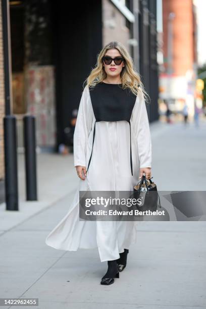 Guest wears black sunglasses, a black breastplate, a white long sleeves long oversized shirt dress, a black shiny leather handbag from Jil Sander,...