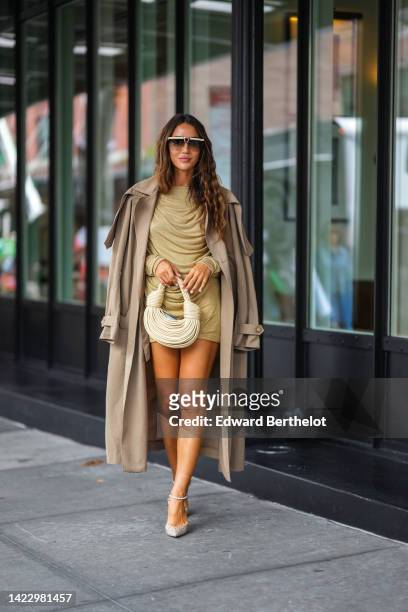 Tamara Kalinic wears beige and brown futurist sunglasses, a gold wrap short dress, a beige long trench coat, a beige matte leather laces handbag, a...