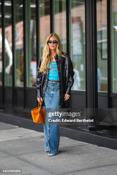 Emili Sindlev wears blue sunglasses, gold earrings, a blue with black and red inscriptions print pattern t-shirt, a black shiny leather oversized...