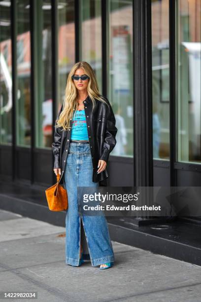 Emili Sindlev wears blue sunglasses, gold earrings, a blue with black and red inscriptions print pattern t-shirt, a black shiny leather oversized...