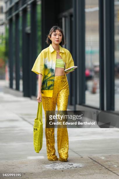 Guest wears gold earrings, gold chain pendants necklaces, a yellow with white and dark green tie and dye print pattern short sleeves shirt, a pale...