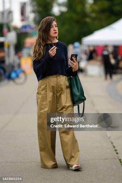 Guest wears a gold long chain pendant necklace, a navy blue wool pullover, gold rings, a green shiny leather Telfar handbag, brown beige wide legs...