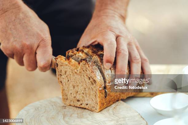 外のテーブルでパンを切る年配の男性の手のクローズアップ - loaf of bread ストックフォトと画像