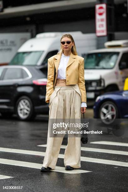 Guest wears black cat eyes sunglasses, a white cropped shirt, a pale yellow linen cropped blazer jacket, beige pleated large pants, a black shiny...