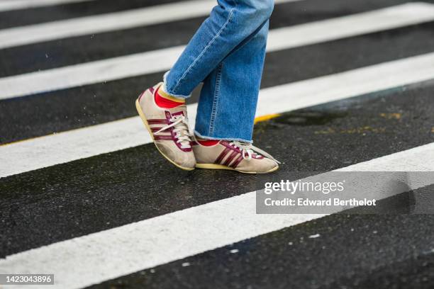 Guest wears blue denim faded ripped jeans pants, beige suede and burgundy shiny varnished leather logo sneakers from Adidas, outside Khaite, during...