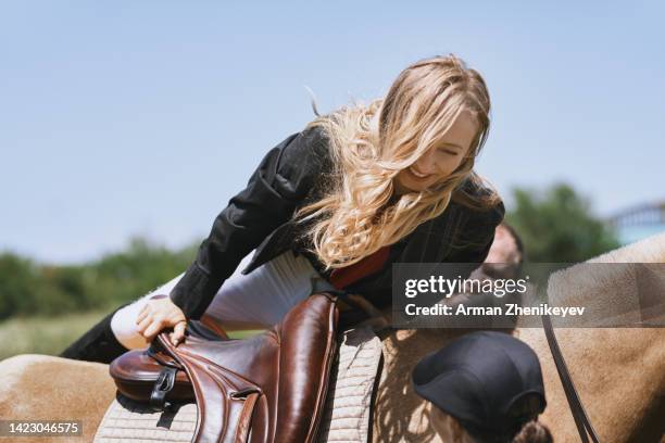 happy blond woman getting on her horse for a ride in a non-urban environment. friends are stabilizing horse and assisting her to climb - enable horse stock-fotos und bilder