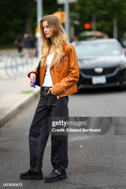 Guest wears a white tank-top, a camel shiny leather zipper jacket, a black shiny leather shoulder bag, black denim wide legs pants, black nylon shoes...