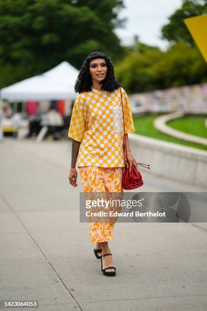 Guest wears a white and pale orange checkered print pattern oversized t-shirt, a red shiny leather shoulder bag, orange / yellow / pink flower print...