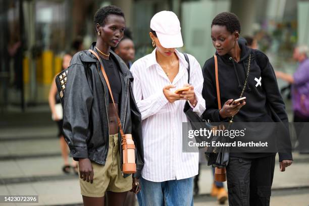 Guest wears gold earrings, a black cropped top from Courreges, a black shiny leather coat, khaki shorts, a brown shiny leather water bottle holder, a...