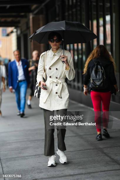 Guest wears a black umbrella, black sunglasses, a pale yellow turtleneck pullover, a beige buttoned / belted trench coat, gray flared pants, white...