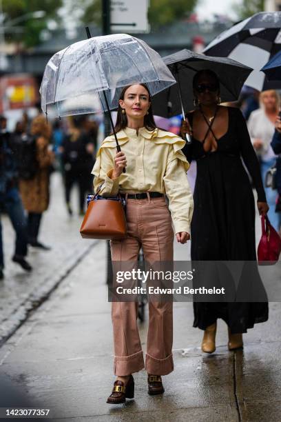Guest wears a pale yellow ruffled shoulder blouse, a brown shiny leather handbag from Staud, a black shiny leather belt, brown denim wide legs pants,...