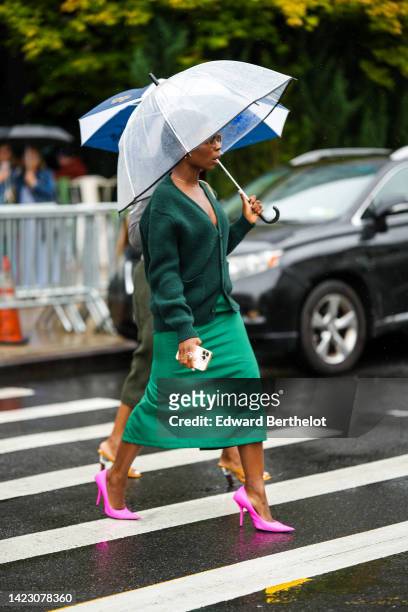 Guest wears sunglasses, a gold necklace, a dark green wool cardigan, a green long skirt, neon pink shiny silk pointed pumps heels shoes, rings ,...