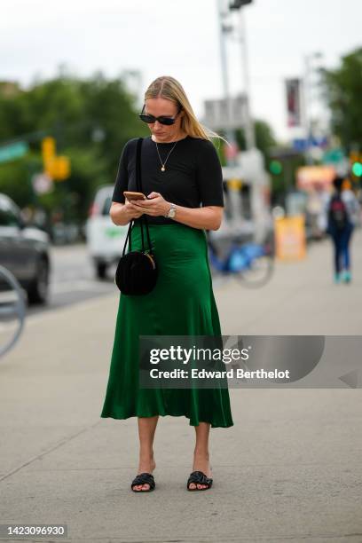 Guest wears black sunglasses, a gold chain pendant necklace, a black short sleeves t-shirt, a dark green midi silk skirt, a black shiny velvet circle...