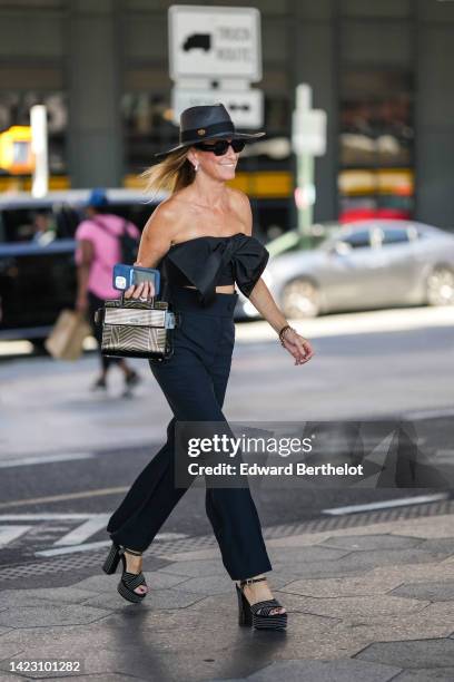 Guest wears a dark gray wicker hat, black sunglasses, diamonds earrings, a black shoulder-off / large knot cropped top, a black with small beige...