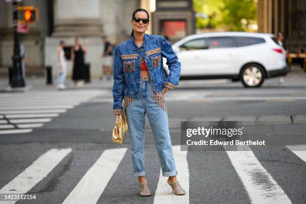 Guest wears black sunglasses, gold earrings, a blue denim with embroidered fringed jewels detail jacket, a red strappy top, blue faded denim large...