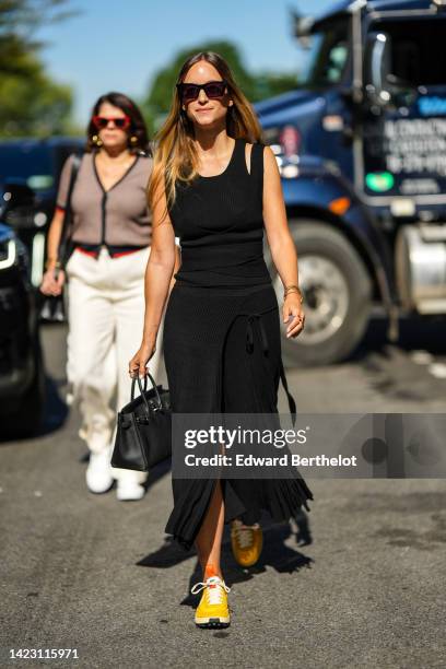 Guest wears black sunglasses, gold earrings, a black ribbed tank-top, a black long skirt, a black shiny leather Birkin handbag from Hermes, yellow...