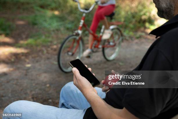 man sitting with a mobile phone in his hand, along a footpath with a child cycling past on a red bicycle in the background. - children screen stock-fotos und bilder