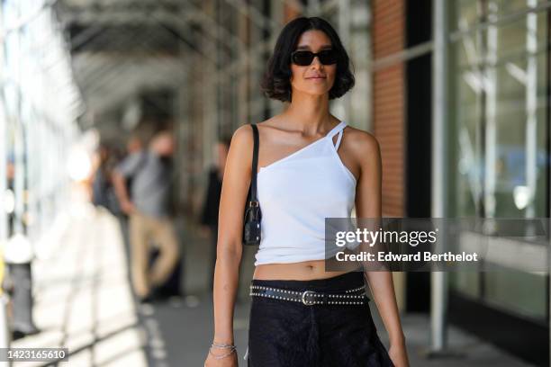 Guest wears black sunglasses, a black shiny leather shoulder bag, a white tank-top, a black shiny leather nailed / studded belt, a black midi skirt,...