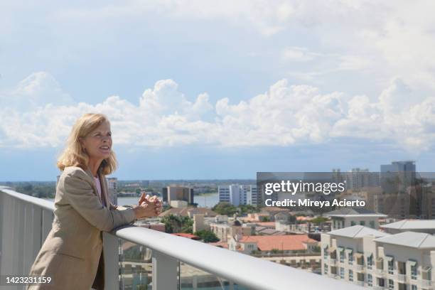 a mature caucasian woman with blond shoulder length hair stands on a balcony of a high rise building overlooking the downtown district of tropical climate city wearing a light beige jacket. - looking over balcony stock pictures, royalty-free photos & images