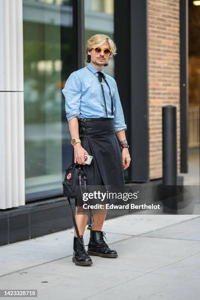 Guest wears yellow sunglasses, a pale blue shirt, a black velvet ribbon tie, a silver large chain necklace, a beige watch, a black wrap knees skirt,...