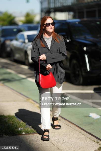 Guest wears black sunglasses, a gray t-shirt, a black oversized jacket, a red shiny varnished leather Cleo handbag from Prada, white denim pants,...