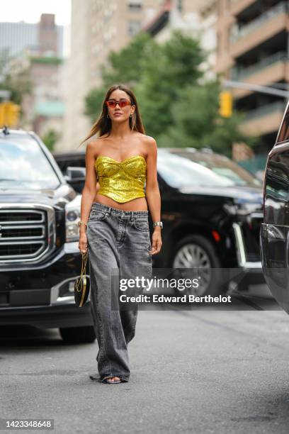 Tamara Kalinic wears red sunglasses, gold earrings, a gold shiny sequined heart shoulder-neck / cropped top, a black shiny leather zipper handbag in...