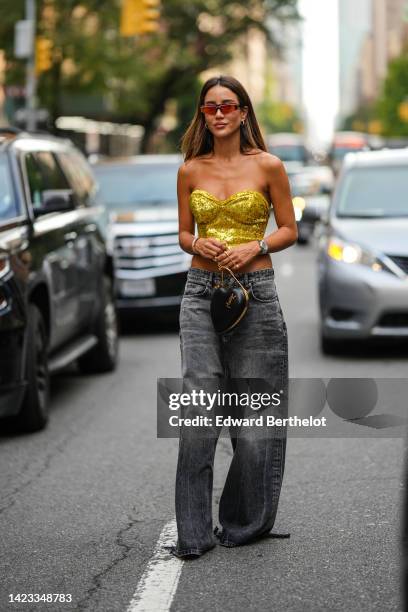 Tamara Kalinic wears red sunglasses, gold earrings, a gold shiny sequined heart shoulder-neck / cropped top, a black shiny leather zipper handbag in...