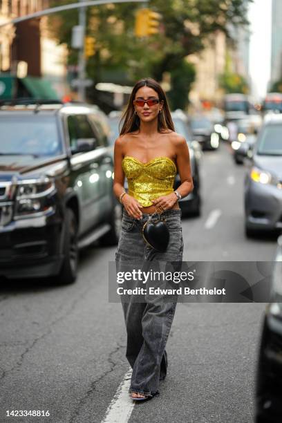 Tamara Kalinic wears red sunglasses, gold earrings, a gold shiny sequined heart shoulder-neck / cropped top, a black shiny leather zipper handbag in...
