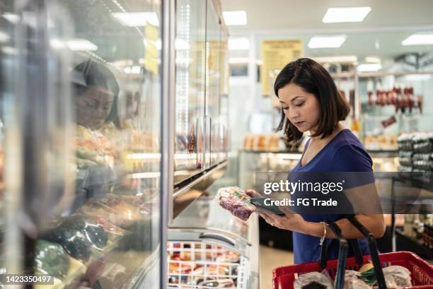 mid adult woman using the mobile phone buying groceries in a supermarket - supermarket freezer stock pictures, royalty-free photos & images