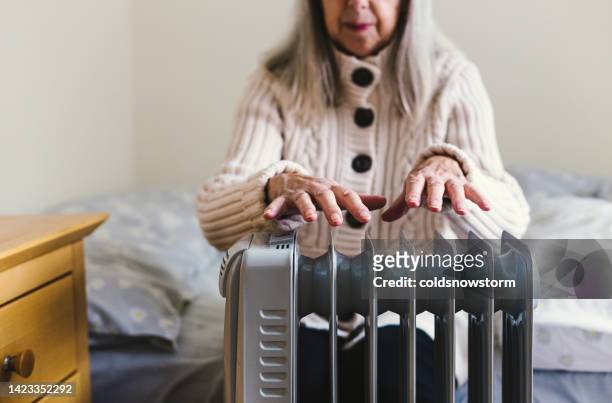senior woman warming her hands over electric heater at home - warme kleding stockfoto's en -beelden