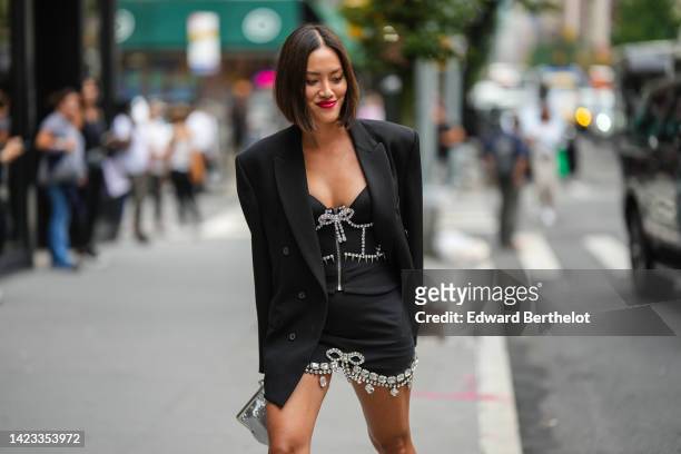 Tiffany Hsu wears a black blazer jacket, a black with embroidered crystal borders V-neck / short dress, a silver shiny leather clutch , outside Area,...