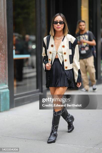 Aimee Song wears black square sunglasses, a silver necklace, a beige and black square print pattern buttoned jacket, a beige gilet, a black pleated /...