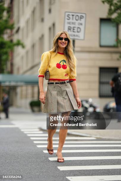 Emili Sindlev wears black sunglasses, gold earrings, a pale yellow with red and green cherry print pattern short sleeves wool pullover, a black shiny...