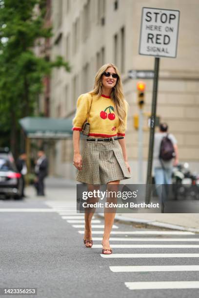 Emili Sindlev wears black sunglasses, gold earrings, a pale yellow with red and green cherry print pattern short sleeves wool pullover, a black shiny...