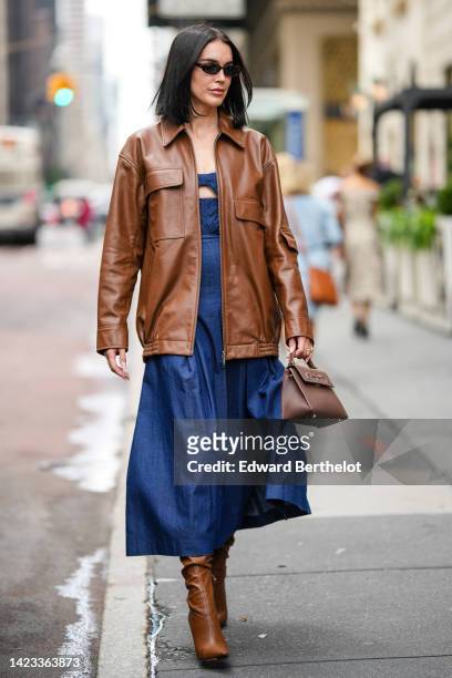 Brittany Xavier wears black sunglasses, a dark brown shiny leather zipper jacket, a navy blue denim long dress, a dark brown shiny leather handbag,...