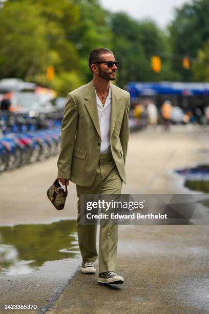 Pelayo Diaz wears black sunglasses, a gold chain pendant necklace, a white shirt, a green oversized blazer jacket, matching green suit pants, a beige...